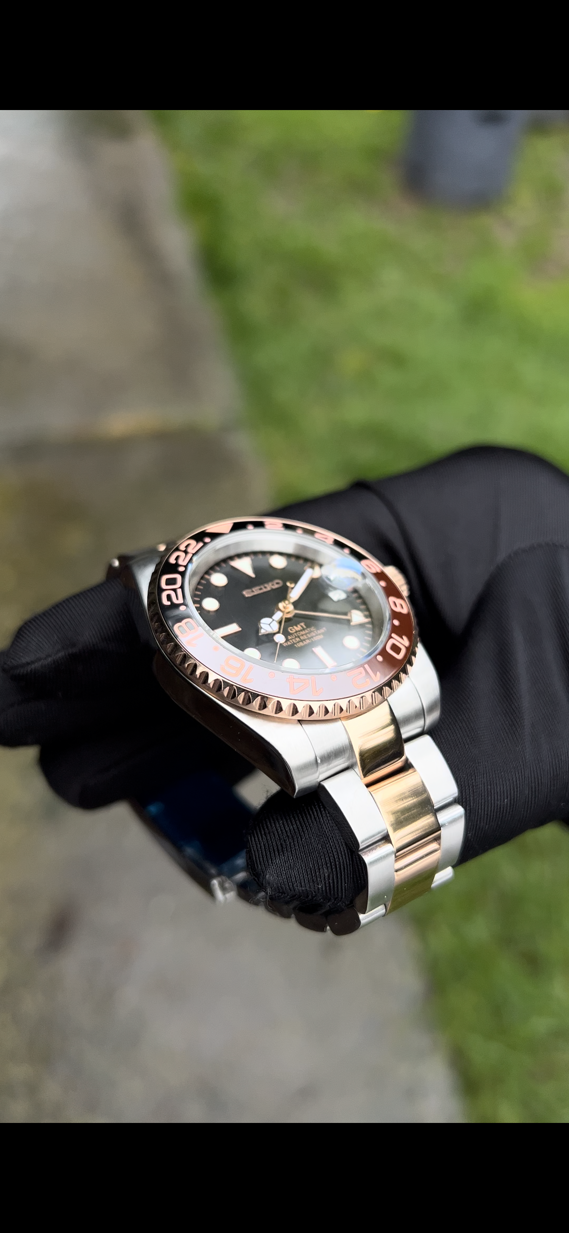 GMT (Rootbeer) Seiko NH34 GMT automatic mod watch. A full watch with a black dial and a black and rose gold bezel on a two tone oyster bracelet held in a glove.