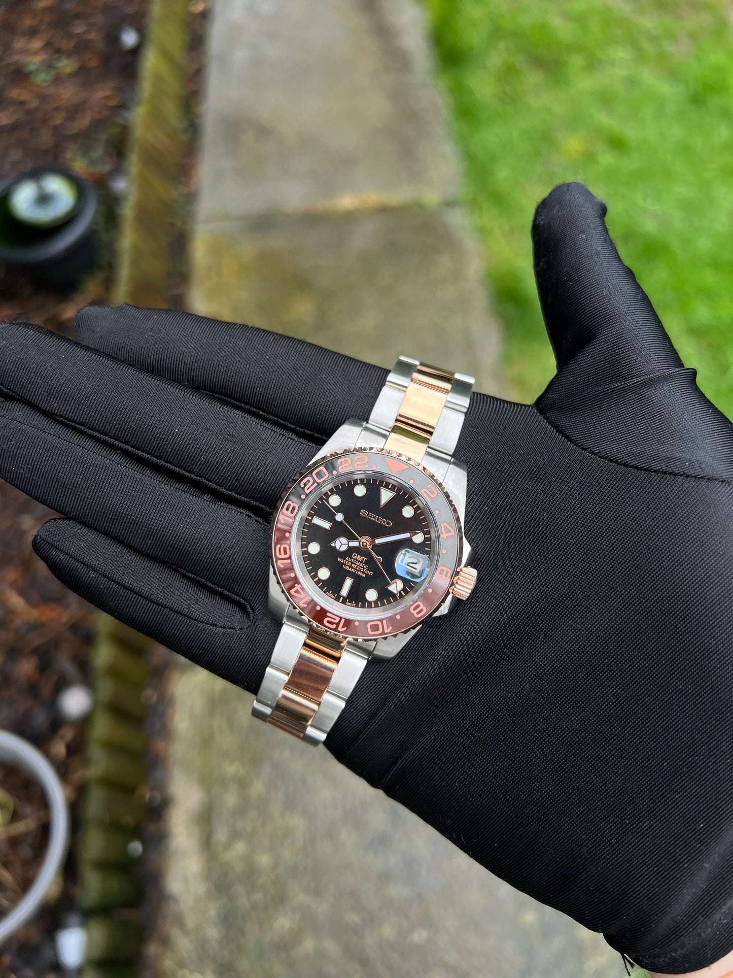 GMT (Rootbeer) Seiko NH34 GMT automatic mod watch. A full watch with a black dial and a black and rose gold bezel on a two tone oyster bracelet held in a glove.