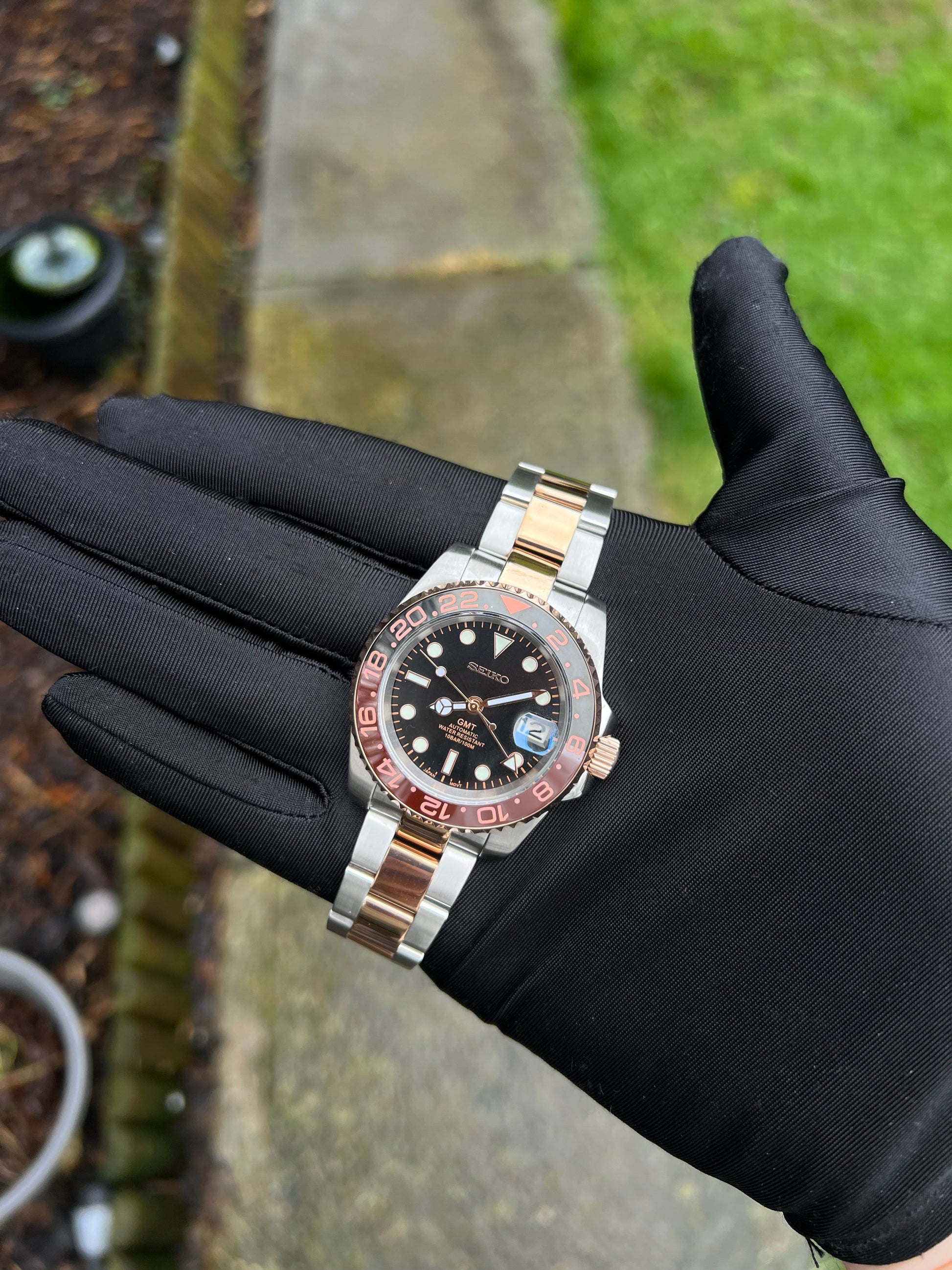 GMT (Rootbeer) Seiko NH34 GMT automatic mod watch. A full watch with a black dial and a black and rose gold bezel on a two tone oyster bracelet held in a glove.