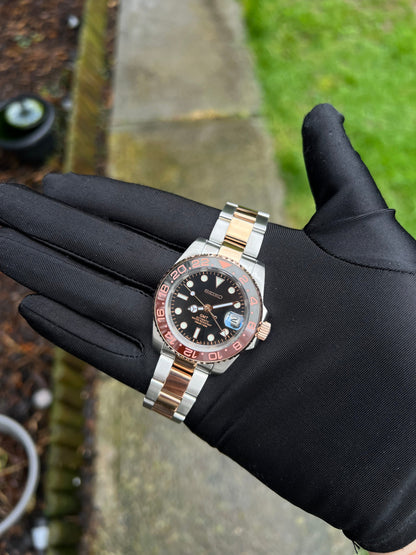 GMT (Rootbeer) Seiko NH34 GMT automatic mod watch. A full watch with a black dial and a black and rose gold bezel on a two tone oyster bracelet held in a glove.