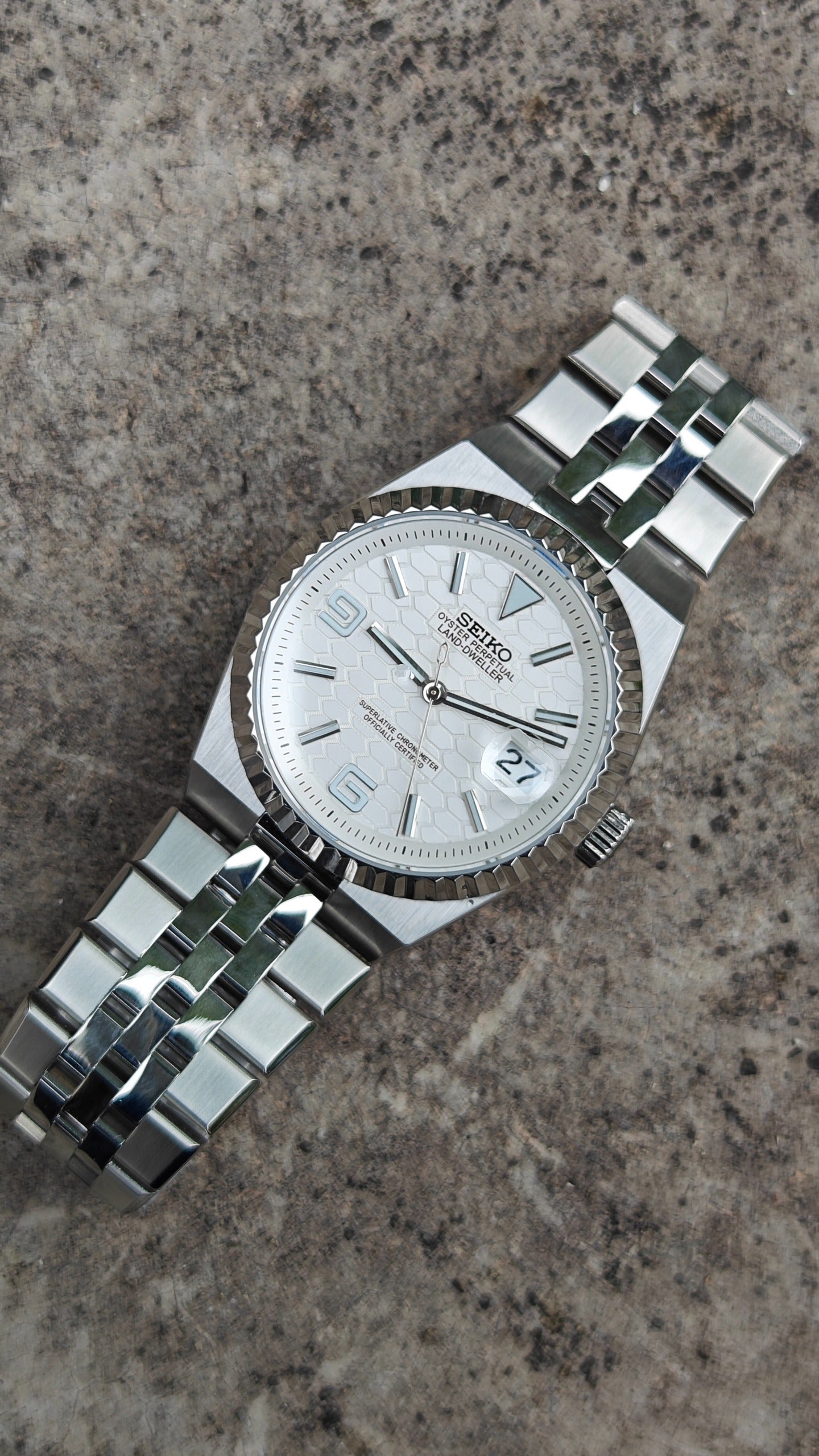 Land-Dweller Seiko NH35 automatic mod watch. A top down view of a white dial watch with a silver fluted bezel and metal link bracelet on stone.