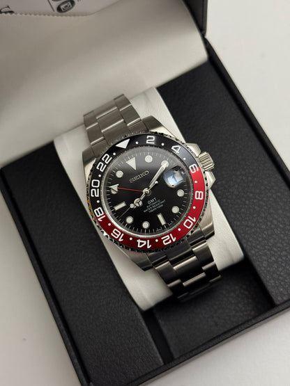GMT (Coke) Seiko NH34 GMT automatic mod watch. A black dial watch with a black and red bezel on a metal link bracelet inside a box in a high angle shot.