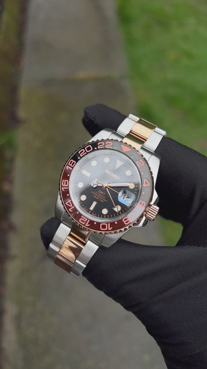 GMT (Rootbeer) Seiko NH34 GMT automatic mod watch. A full watch with a black dial and a black and rose gold bezel on a two tone oyster bracelet held in a glove.