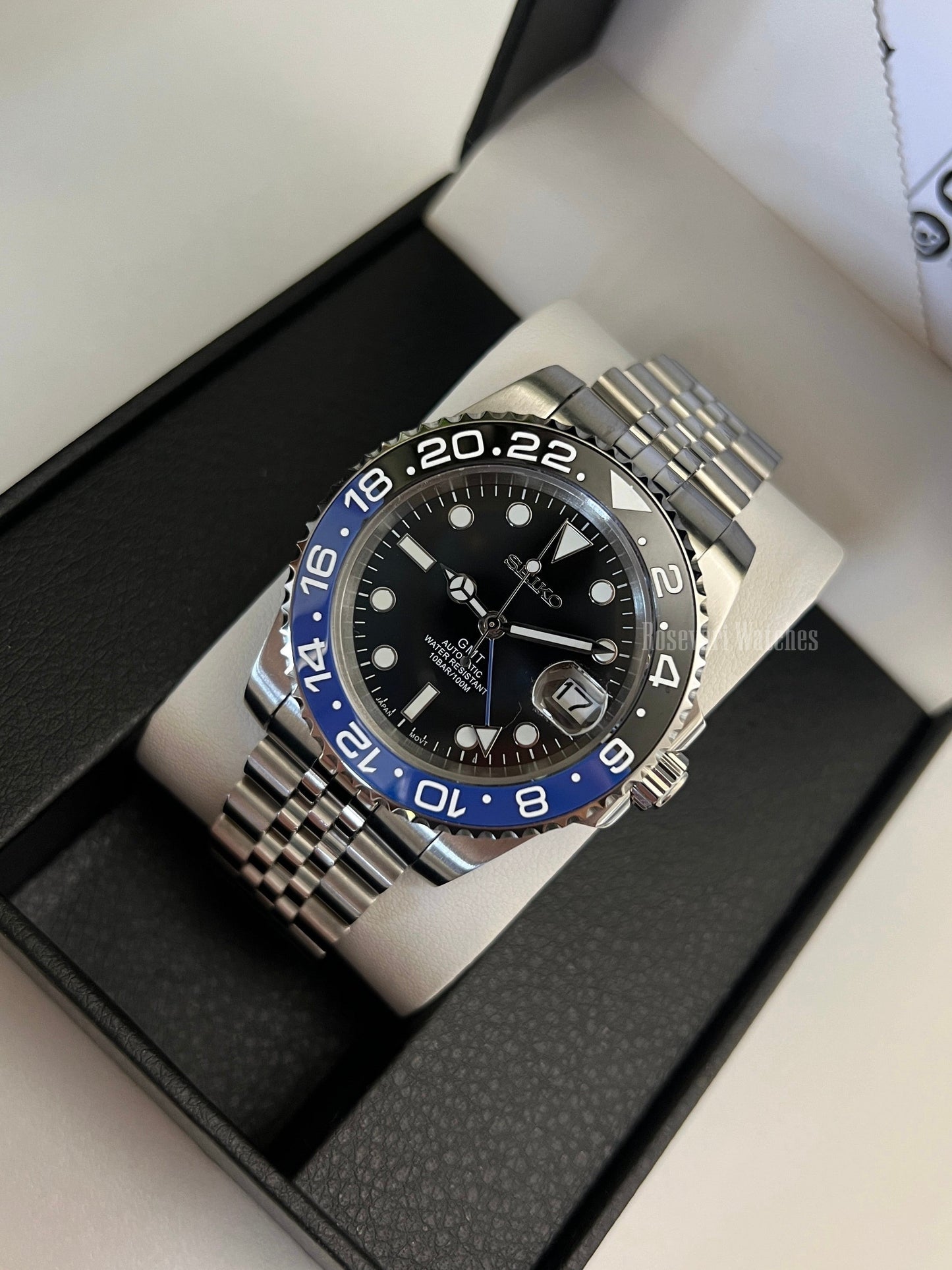 GMT (Batman) Seiko NH34 GMT automatic mod watch. An angled full watch shot in a box with a black dial and blue and black bezel on a silver bracelet.