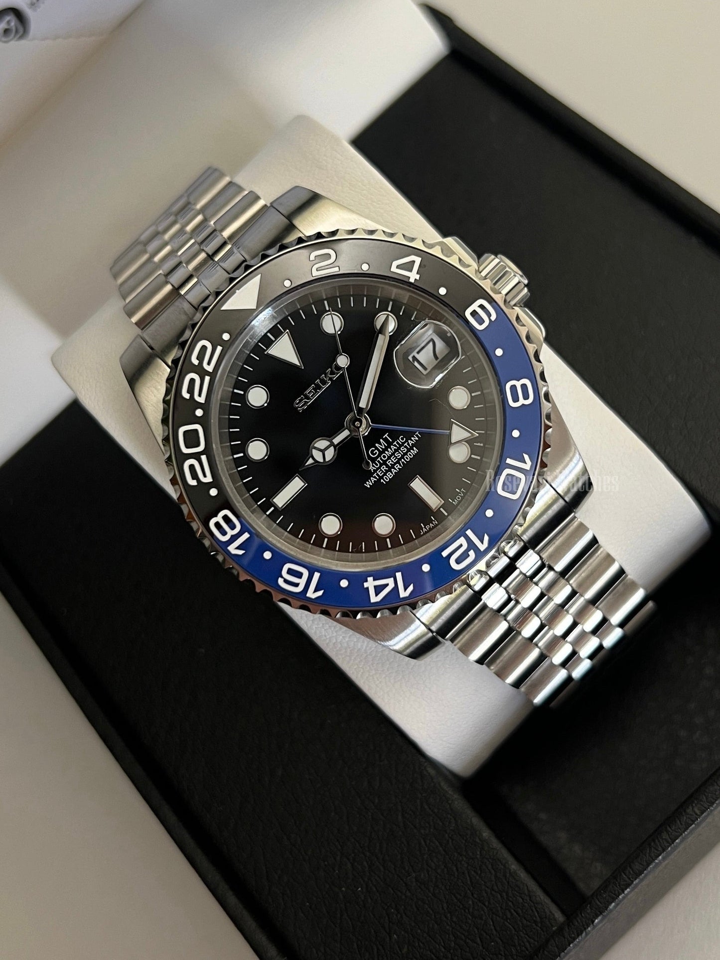 GMT (Batman) Seiko NH34 GMT automatic mod watch. A high angle full watch shot in a box with a black dial and blue and black bezel on a silver bracelet.