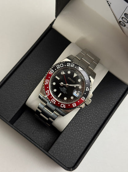 GMT (Coke) Seiko NH34 GMT automatic mod watch. A black dial watch with a black and red bezel on a metal link bracelet inside a box in a high angle shot.
