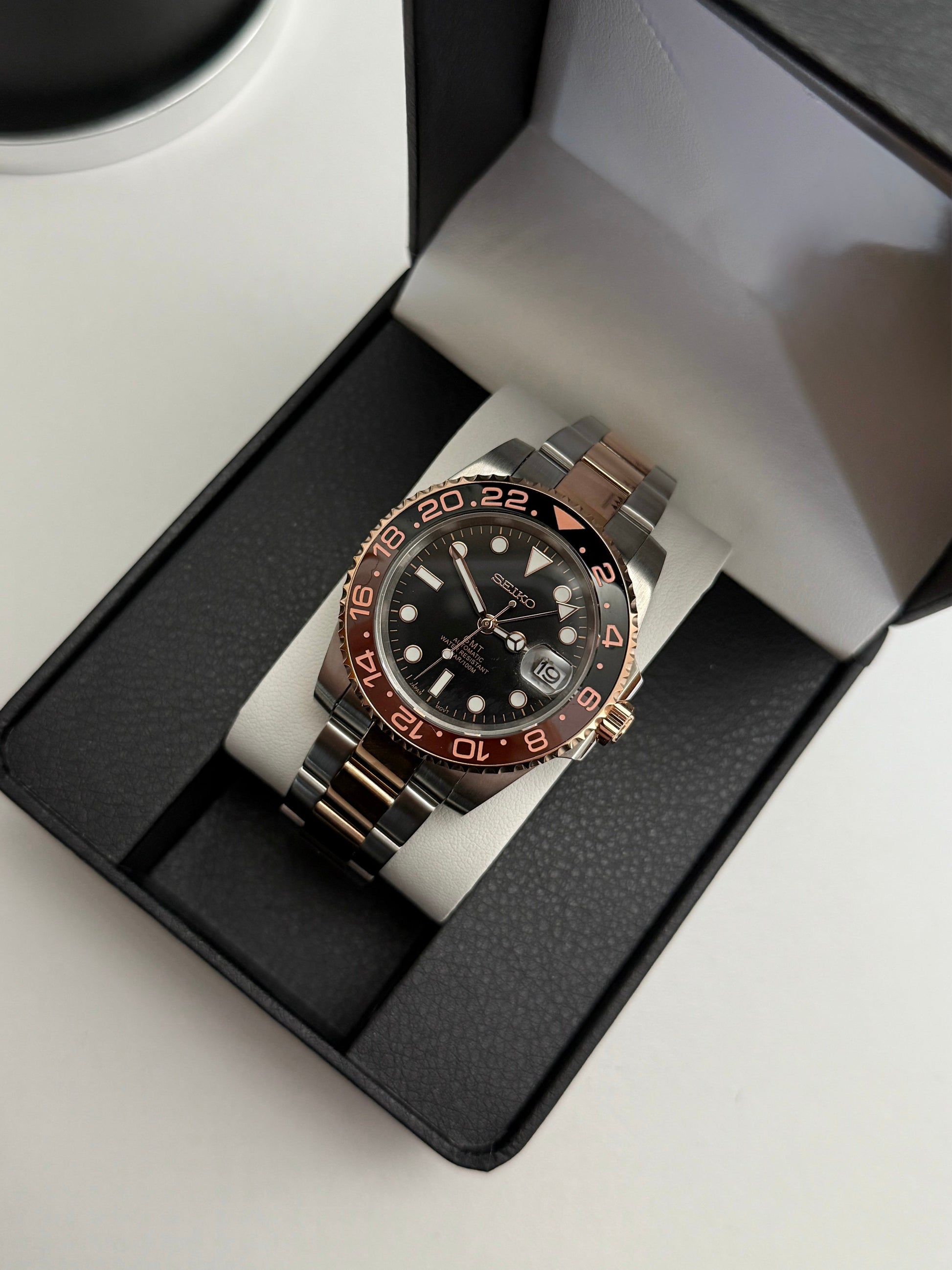 GMT (Rootbeer) Seiko NH34 GMT automatic mod watch. A full watch with a black dial and a black and rose gold bezel on a two tone oyster bracelet in a box.