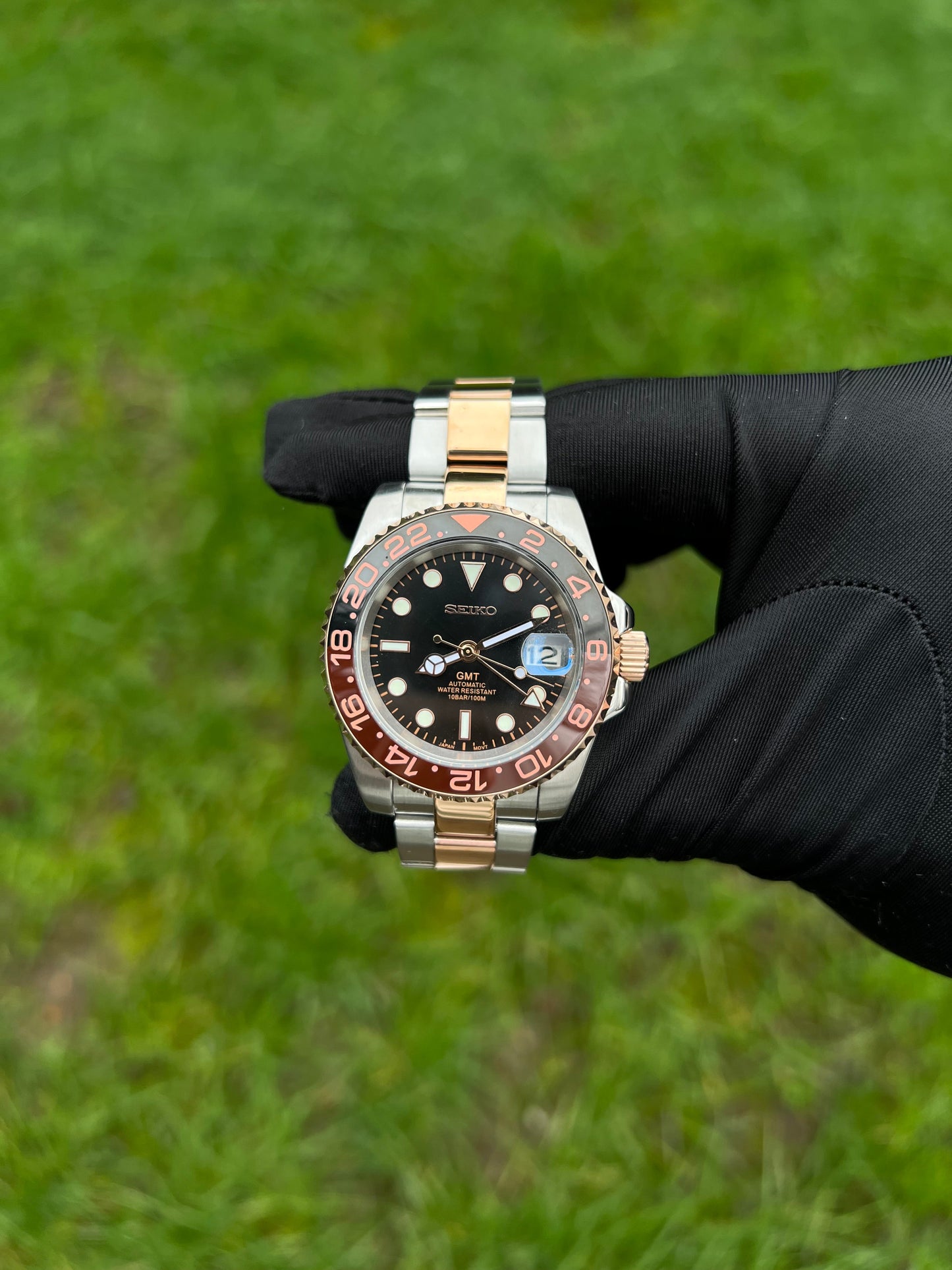 GMT (Rootbeer) Seiko NH34 GMT automatic mod watch. A full watch with a black dial and a black and rose gold bezel on a two tone oyster bracelet held in a glove.