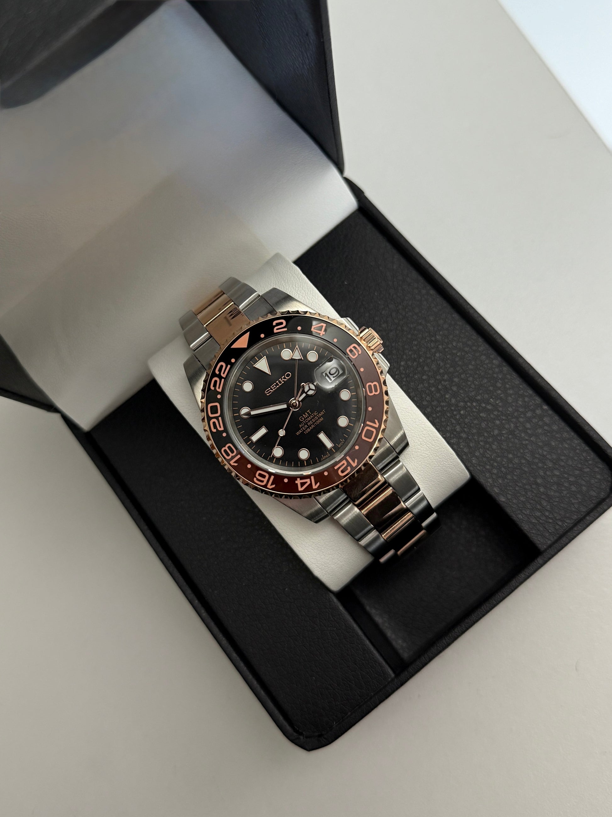 GMT (Rootbeer) Seiko NH34 GMT automatic mod watch. A full watch with a black dial and a black and rose gold bezel on a two tone oyster bracelet in a box.