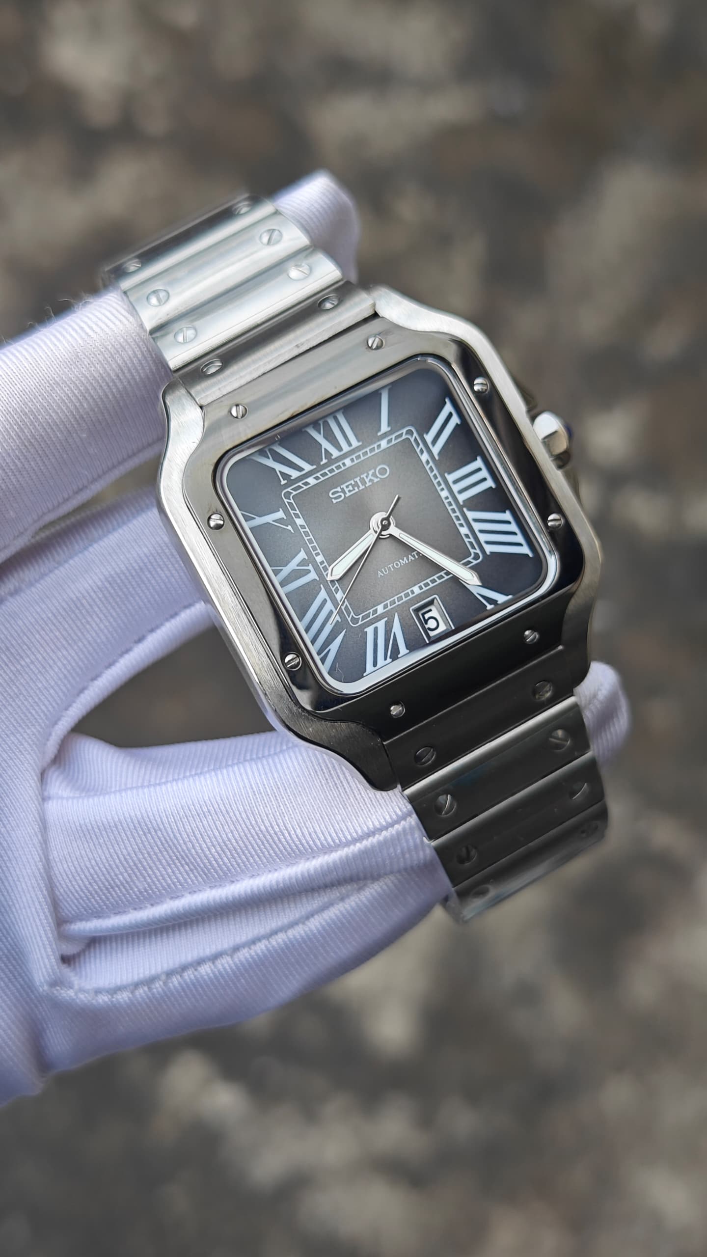 Santos (Graphite) Seiko NH35 automatic mod watch. A full watch shot held by a gloved hand showing a grey dial with a black bezel and a metal link bracelet.